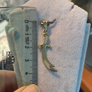 K&ouml;nnte beinhalten: Ein goldfarbener Anh&auml;nger in Form eines gebogenen Schwertes mit einem Griff, der mit kleinen, klaren Edelsteinen verziert ist. Der Anh&auml;nger ist etwa 2,5 cm lang, gemessen mit einem Lineal. Der Anh&auml;nger wird auf einer hellen Oberfl&auml;che pr&auml;sentiert.