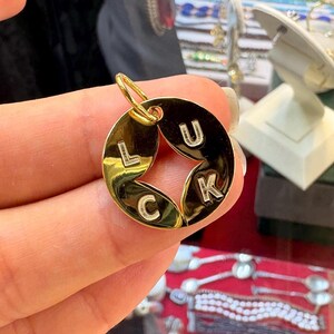 May include: A gold-colored pendant with the word "LUCK" spelled out. The letters are white and are set against a dark background. The pendant has a circular shape with a star-shaped cutout in the center. A small loop is at the top for a chain.