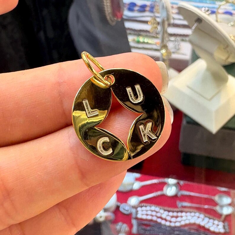 May include: A gold-colored pendant with the word "LUCK" spelled out. The letters are white and are set against a dark background. The pendant has a circular shape with a star-shaped cutout in the center. A small loop is at the top for a chain.