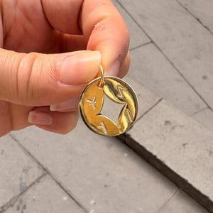May include: A gold-colored pendant with a circular design. The pendant features a central star-shaped cutout and stylized symbols. The pendant is held up by a hand.