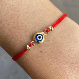 Puede incluir: Una pulsera de hilo rojo con un dije dorado que presenta un diseño de ojo azul y blanco. La pulsera tiene cuentas doradas a cada lado del dije. La pulsera se usa en la muñeca.
