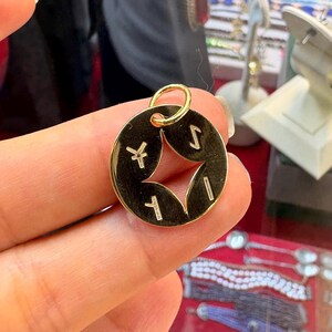 May include: A gold-colored pendant with a circular shape and a star-shaped cutout in the center. The pendant has four symbols around the star and a small loop at the top for a chain. The pendant is held in a person's hand.