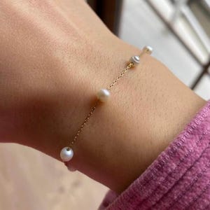 Puede incluir: Una delicada pulsera dorada con pequeñas perlas blancas redondas. Las perlas están espaciadas a lo largo de una fina cadena dorada. La pulsera se muestra en una muñeca, con una manga rosa en el fondo.