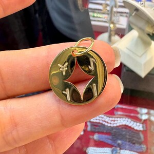 May include: A gold-colored pendant with a circular design. The pendant features a star-shaped cutout in the center and four symbols around the edges. A small loop is at the top for attaching to a chain or bracelet.