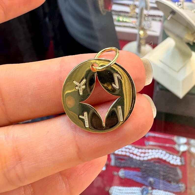 May include: A gold-colored pendant with a circular design. The pendant features a star-shaped cutout in the center and four symbols around the edges. A small loop is at the top for attaching to a chain or bracelet.