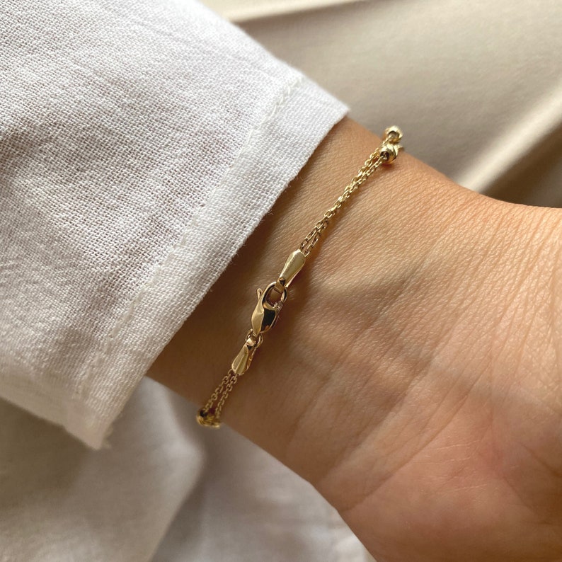 Puede incluir: Una delicada pulsera dorada con un dise&ntilde;o de doble cadena. La pulsera presenta peque&ntilde;as cuentas redondas y un cierre seguro. La joya se muestra en una mu&ntilde;eca, con una camisa blanca en el fondo.