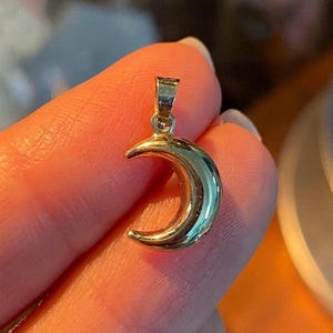 Puede incluir: Un colgante de luna creciente de color dorado. El colgante tiene una superficie lisa y brillante, con una forma curvada. Cuelga de una fina cadena. El diseño es simple y elegante, adecuado para el uso diario. Mide aproximadamente 1,5 cm.
