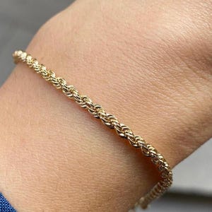Puede incluir: Un brazalete de cadena de cuerda de color dorado. El brazalete está hecho de eslabones tejidos apretados, creando una superficie texturizada. El brazalete se muestra en la muñeca de una persona.