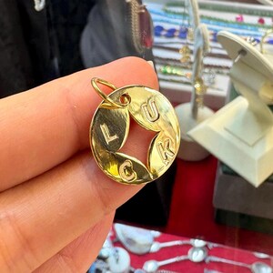 May include: A gold-colored circular pendant with the word "LUCK" spelled out. The letters are cut out of the center, forming a star shape. The pendant has a small loop at the top for a chain.