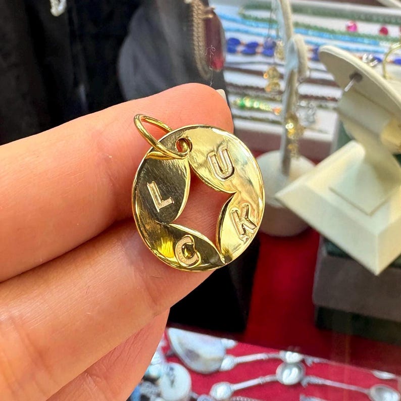 May include: A gold-colored circular pendant with the word "LUCK" spelled out. The letters are cut out of the center, forming a star shape. The pendant has a small loop at the top for a chain.