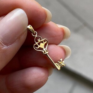 May include: A gold-colored key pendant with an ornate design. The key has a decorative top with a diamond-shaped accent and a detailed key bit. The pendant is held up by a small loop for a chain.