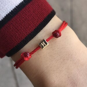 Puede incluir: Pulsera de hilo rojo con un dije cuadrado dorado con la letra "H". La pulsera tiene dos nudos decorativos y se lleva en la muñeca. La manga de una camisa a rayas rojas y blancas es visible en el fondo.