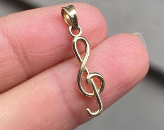 May include: A gold-toned metal pendant shaped like a treble clef musical symbol. The pendant features a smooth, polished surface with a small integrated loop at the top for attachment to a chain or cord, displaying a classic musical notation design.