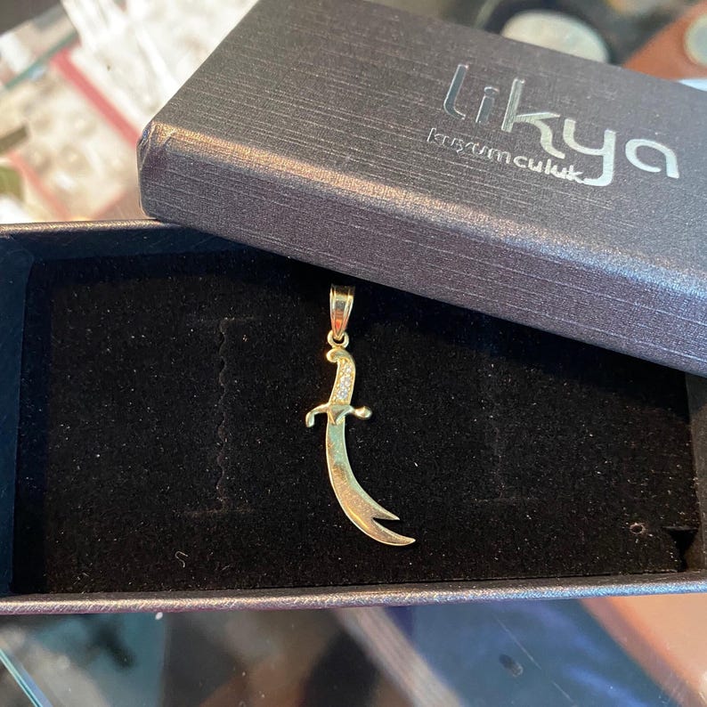 K&ouml;nnte beinhalten: Goldfarbener Anh&auml;nger in Form eines gebogenen Schwertes mit einem Griff, der mit kleinen, klaren Steinen besetzt ist. Der Anh&auml;nger wird in einer schwarzen Schmuckschatulle mit dem Markennamen "Likya" pr&auml;sentiert.
