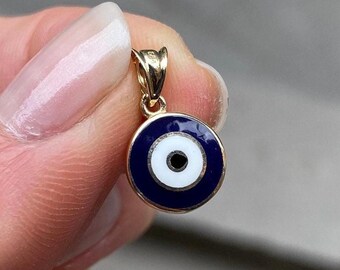 Pode incluir: Pingente dourado com um design de olho grego esmaltado em azul e branco. O charme redondo tem uma borda azul escura, um centro branco e um ponto preto no meio. O pingente é preso por um laço dourado.