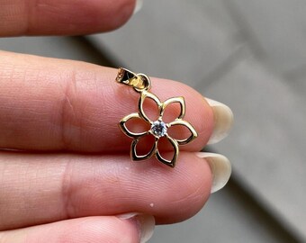 May include: A gold pendant shaped like a six-petaled flower with openwork petals and a single clear, round-cut gemstone set in the center. The pendant features a small, polished gold bail at the top for attachment to a chain or necklace.
