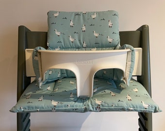 May include: A white high chair with a light blue cushion set featuring a pattern of white geese. The high chair has a white tray and a gray frame. The cushion set includes a seat cushion, back cushion, and side cushions.
