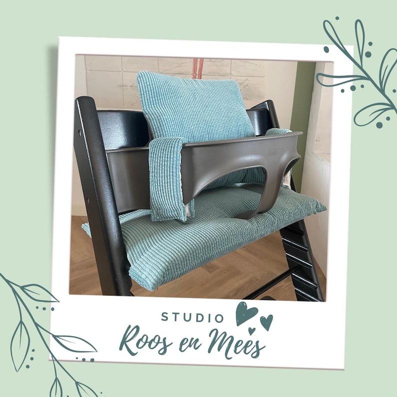 May include: A black high chair with a teal blue cushion and a teal blue pillow. The chair has a tray and a safety harness. The chair is in a room with white tile walls. The text "STUDIO Roos en Mees" is visible in the image.