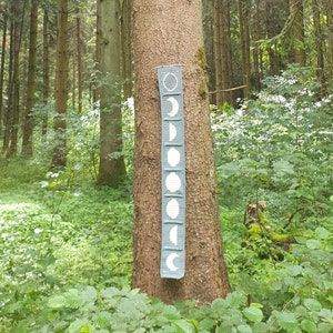 Peut inclure: Une tenture murale en tissu gris avec des phases de lune blanches imprimées dessus. La tenture est attachée au tronc d'un arbre dans un environnement forestier.