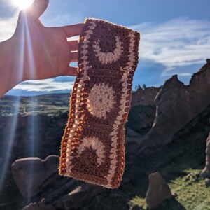 Puede incluir: Una diadema de ganchillo marrón y crema con un diseño de espiral repetido. La diadema se sostiene contra un cielo azul y un paisaje montañoso. El trabajo de ganchillo parece hecho a mano.
