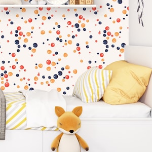 May include: A white wall with a repeating pattern of orange, red, and blue circles. A white shelf with a small plant, a toy, and a wooden sign with the text "Old" is mounted on the wall. A white bed with a gray knitted blanket and yellow and white striped pillows is in front of the wall.