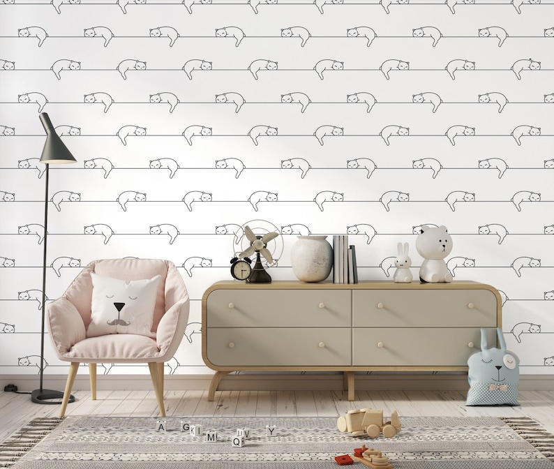 Puede incluir: Papel tapiz blanco con un patr&oacute;n repetitivo de dibujos de l&iacute;nea negra de gatos durmiendo en una l&iacute;nea. Un tocador gris claro con cuatro cajones est&aacute; en primer plano. Un sill&oacute;n rosa con una almohada blanca est&aacute; frente al tocador. Un peque&ntilde;o cami&oacute;n de juguete de madera est&aacute; en el suelo frente al tocador.