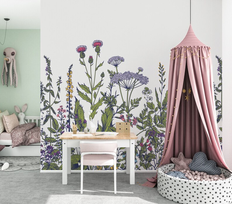 May include: A white wall with a floral mural featuring a variety of green plants and flowers in shades of pink, purple, and yellow. A pink canopy bed with gold stars hangs in the corner of the room. A white desk with a pink chair sits in front of the mural.