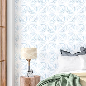May include: A bedroom scene featuring a light blue and white patterned wallpaper. A wooden bedside table holds a lamp with a beige shade and a small alarm clock. White pillows and a green blanket are on the bed.