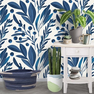 May include: A white wall with a blue floral wallpaper pattern. A white side table with a round top and a lower shelf. A potted plant with green leaves in a white planter with a green and white design. A blue and white rope basket. A potted plant with green leaves in a gray planter. A white mug with a blue and white pattern.