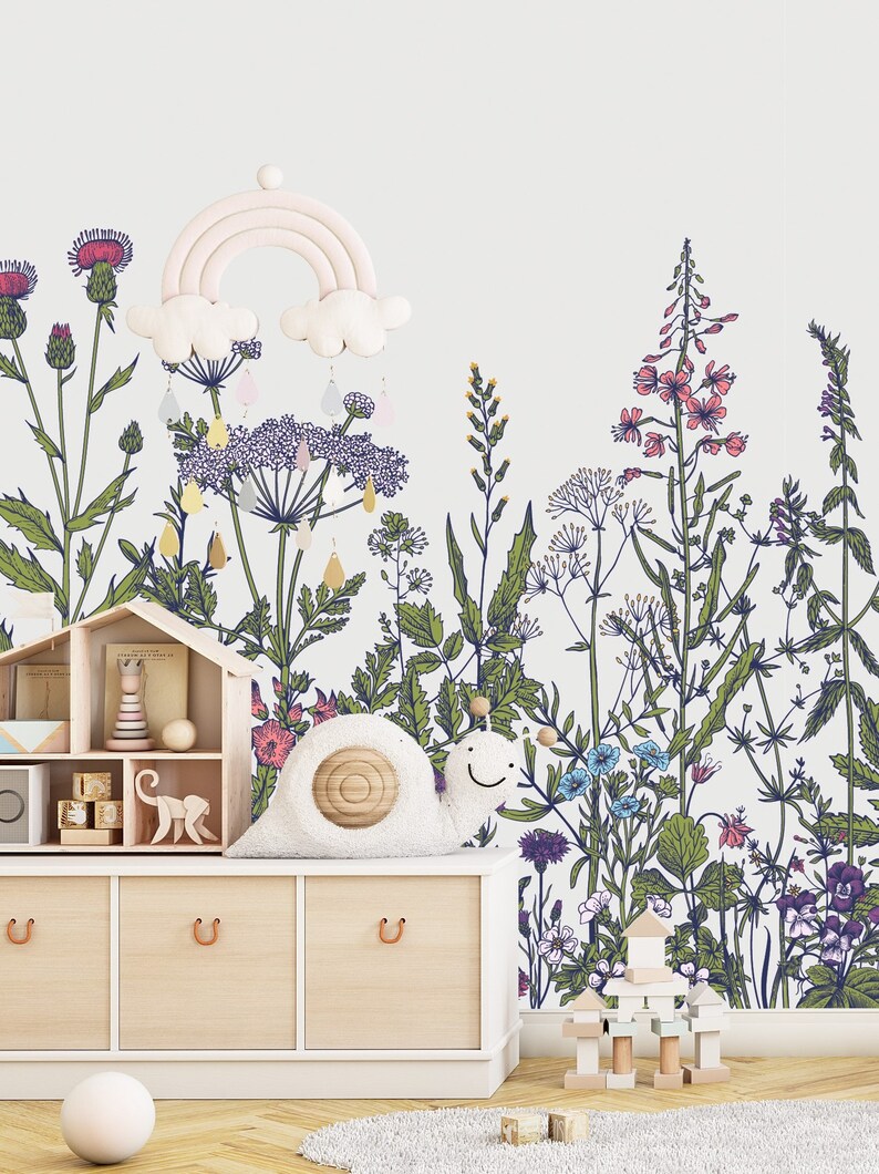 May include: A white wall with a floral wallpaper design featuring a variety of flowers and plants in shades of green, blue, pink, and purple. A white wooden shelf with a toy house, wooden blocks, and a stuffed snail sits in front of the wall.
