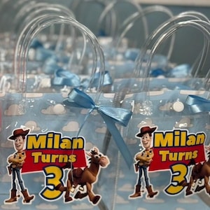 May include: Clear plastic party favor bags with blue ribbon bows. Each bag features a cartoon image of Woody and a horse, with the text "Milan Turns 3" in yellow and red. The bags have clear plastic handles.