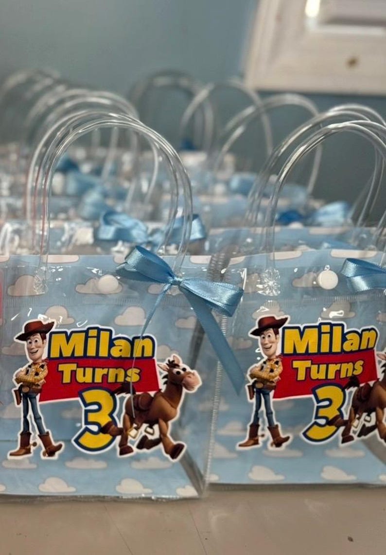May include: Clear plastic party favor bags with blue ribbon bows. Each bag features a cartoon image of Woody and a horse, with the text "Milan Turns 3" in yellow and red. The bags have clear plastic handles.