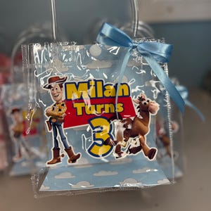 May include: Clear plastic party favor bag with a blue ribbon bow. The bag features a cartoon image of Woody from Toy Story, the text "Milan Turns 3", and a cartoon image of a horse. The background is light blue with white clouds.
