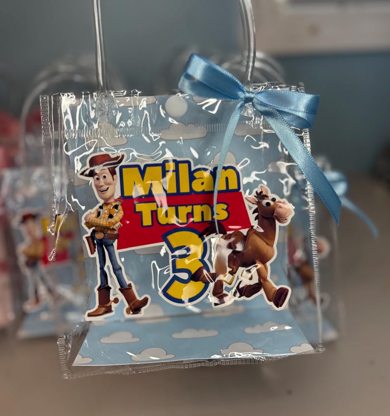 May include: Clear plastic party favor bag with a blue ribbon bow. The bag features a cartoon image of Woody from Toy Story, the text "Milan Turns 3", and a cartoon image of a horse. The background is light blue with white clouds.