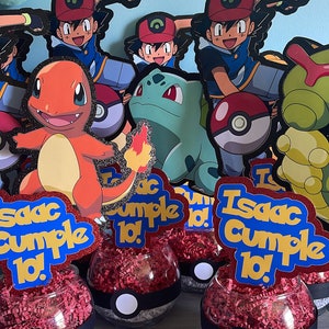 May include: A birthday party centerpiece with a Pokemon theme. The centerpiece features a glass bowl filled with red and white tissue paper, a black and white Pokeball design, and a blue and gold sign that says "Isaac cumple 10!"