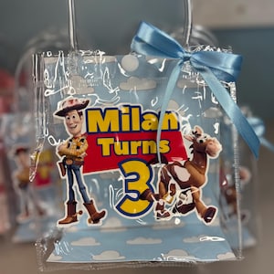May include: Clear plastic party favor bag with a light blue ribbon. The bag features a cartoon image of Woody and a horse, with the text "Milan Turns 3" in yellow and red.