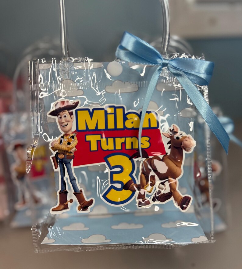 May include: Clear plastic party favor bag with a light blue ribbon. The bag features a cartoon image of Woody and a horse, with the text "Milan Turns 3" in yellow and red.