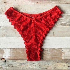 May include: Bright red lace underwear with a scalloped edge and a small bow detail. The lingerie is made of a delicate, patterned lace fabric. The thong-style underwear is displayed on a wooden surface.