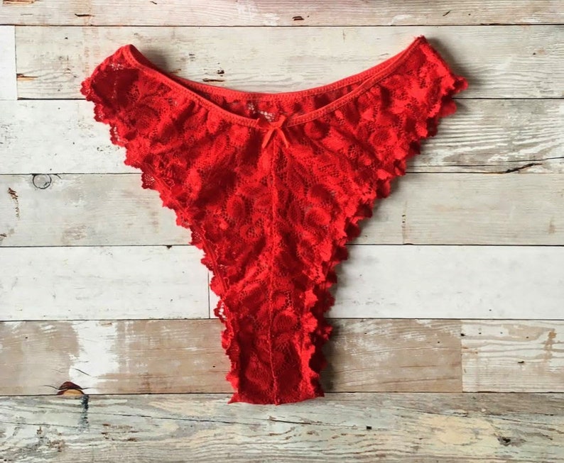 May include: Bright red lace underwear with a scalloped edge and a small bow detail. The lingerie is made of a delicate, patterned lace fabric. The thong-style underwear is displayed on a wooden surface.
