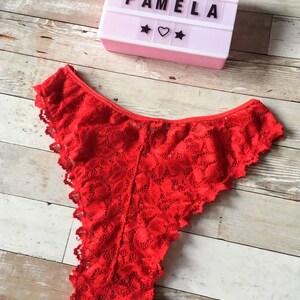 May include: Bright red lace underwear with a scalloped edge. A light-up sign with the text "BOMBI PAMELA" is in the background, along with star and heart symbols. The underwear is made of a delicate, floral-patterned lace.