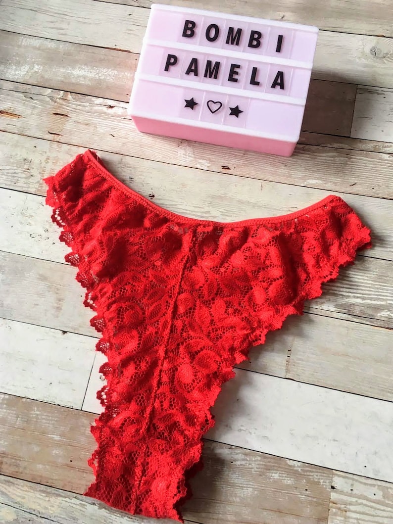 May include: Bright red lace underwear with a scalloped edge. A light-up sign with the text "BOMBI PAMELA" is in the background, along with star and heart symbols. The underwear is made of a delicate, floral-patterned lace.