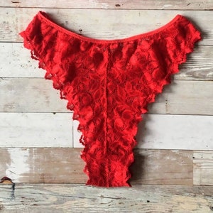 May include: Bright red lace underwear with a scalloped edge. The delicate fabric is patterned with a floral design. The garment is displayed on a light wood surface.