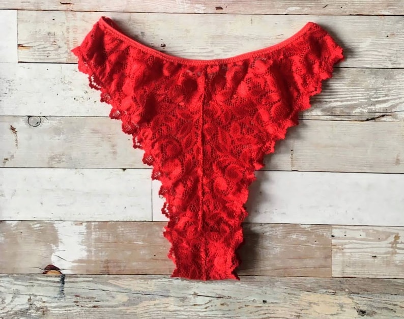 May include: Bright red lace underwear with a scalloped edge. The delicate fabric is patterned with a floral design. The garment is displayed on a light wood surface.