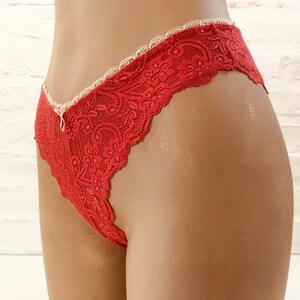 May include: Red lace underwear with a scalloped edge and a small bow detail. The delicate floral pattern and ivory trim create a feminine look. The underwear is displayed on a mannequin.