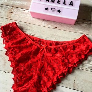 May include: Bright red lace underwear with a scalloped edge. A light pink marquee sign displays the text "BOMBI PAMELA" with star and heart symbols. The items are on a light wood grain surface.