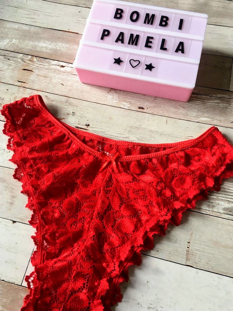 May include: Bright red lace underwear with a scalloped edge. A light pink marquee sign displays the text "BOMBI PAMELA" with star and heart symbols. The items are on a light wood grain surface.