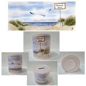 Könnte beinhalten: Weiße Keramik-Sparbüchse mit einer Strandszene und einem Schild mit der Aufschrift "Urlaubs-kasse". Die Szene zeigt eine Möwe, die über dem Meer fliegt, und einen Sandstrand mit Dünen.