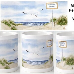 Può includere: Tazza in ceramica bianca con un'illustrazione ad acquerello di una scena di spiaggia. La scena mostra una spiaggia sabbiosa, un oceano blu e un gabbiano che vola sopra. Un cartello recita "Wunschort Strand".
