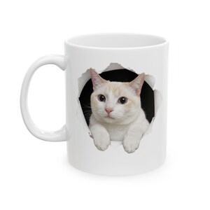 May include: A white ceramic mug featuring a graphic of a cream and white cat with blue eyes peering through a circular, torn-paper effect. The cat is positioned in the center, with its front paws resting on the bottom edge of the black hole.