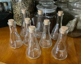 May include: Eight clear glass bottles with cork stoppers. The bottles are arranged in a row on a wooden surface.